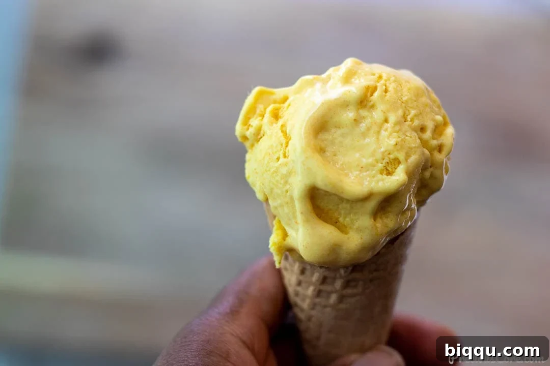 A hand holding a scoop of golden mango ice cream in a crisp waffle cone, against a blurred tropical background.