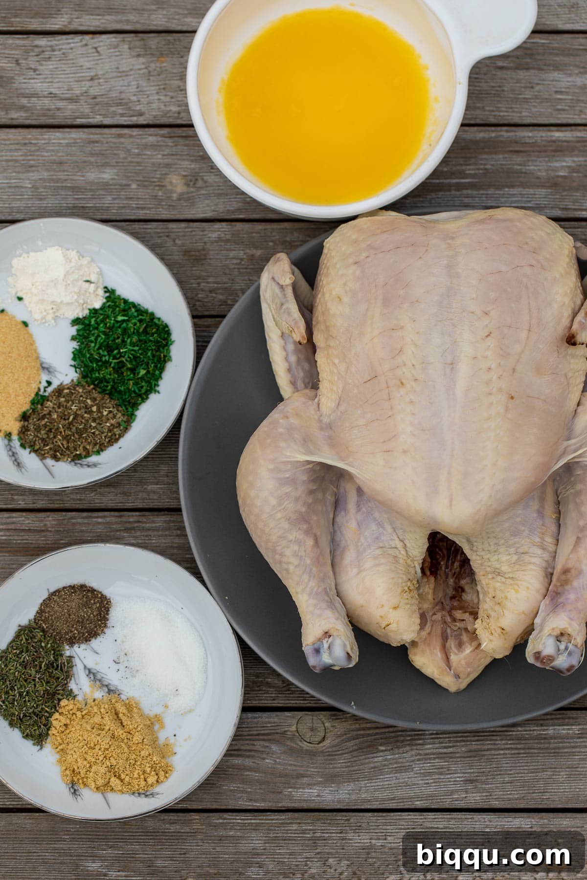 A whole chicken, an assortment of spices, and fresh herbs laid out on a rustic wooden board, ready for preparation.
