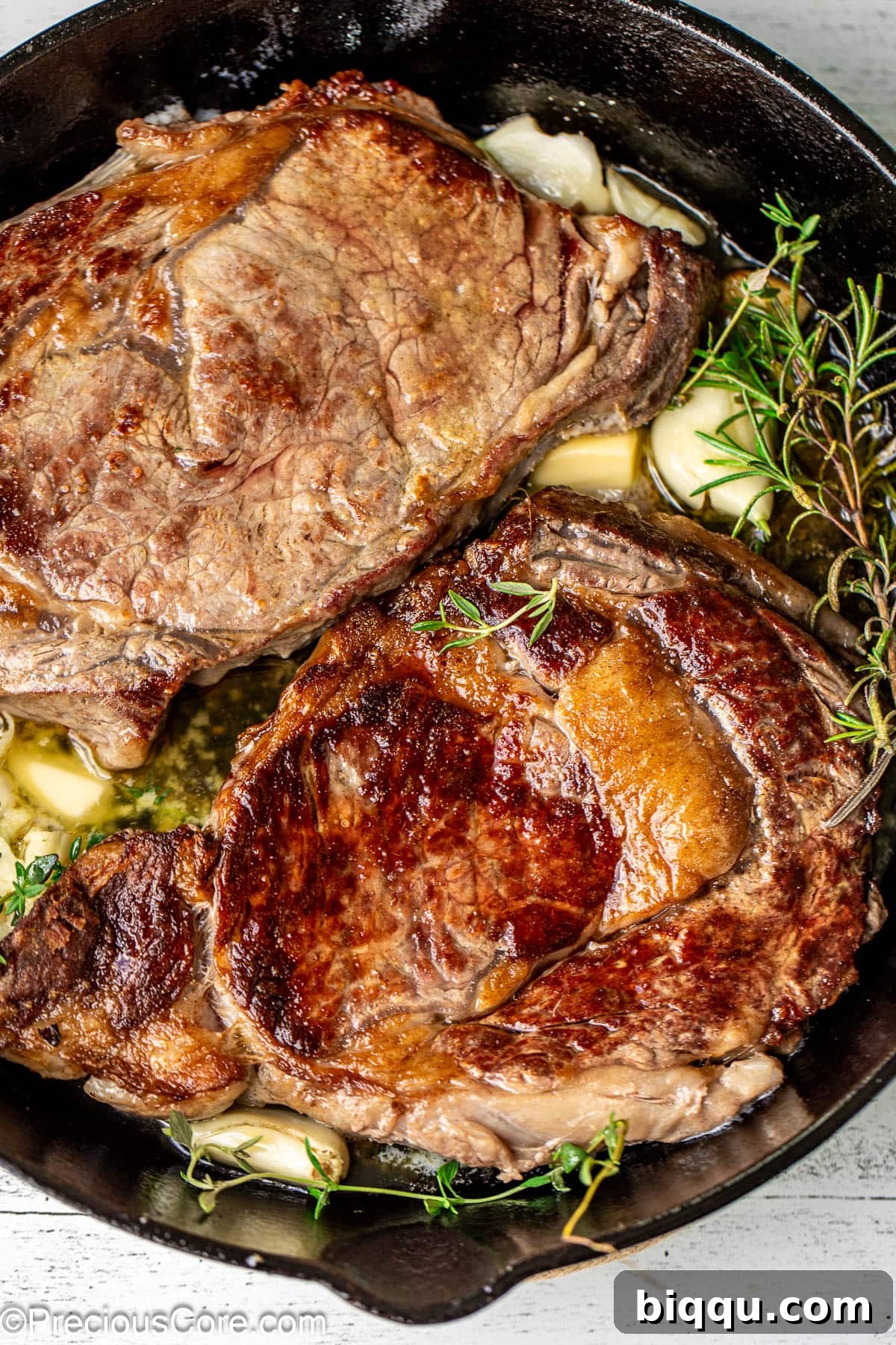 Seared Ribeye Perfection 2 Two perfectly seared ribeye steaks in a cast iron skillet, garnished with fresh rosemary, thyme, and garlic cloves.