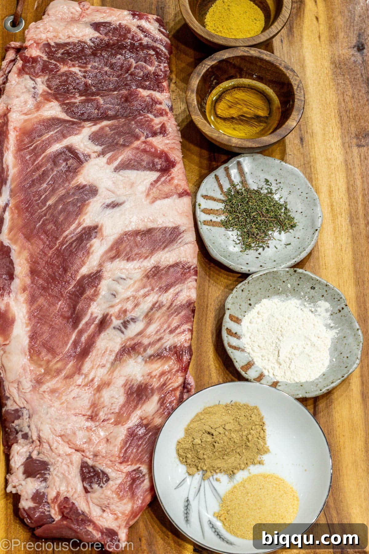 Raw spare ribs laid out with a variety of spices and a salt shaker on a wooden cutting board, ready for preparation.