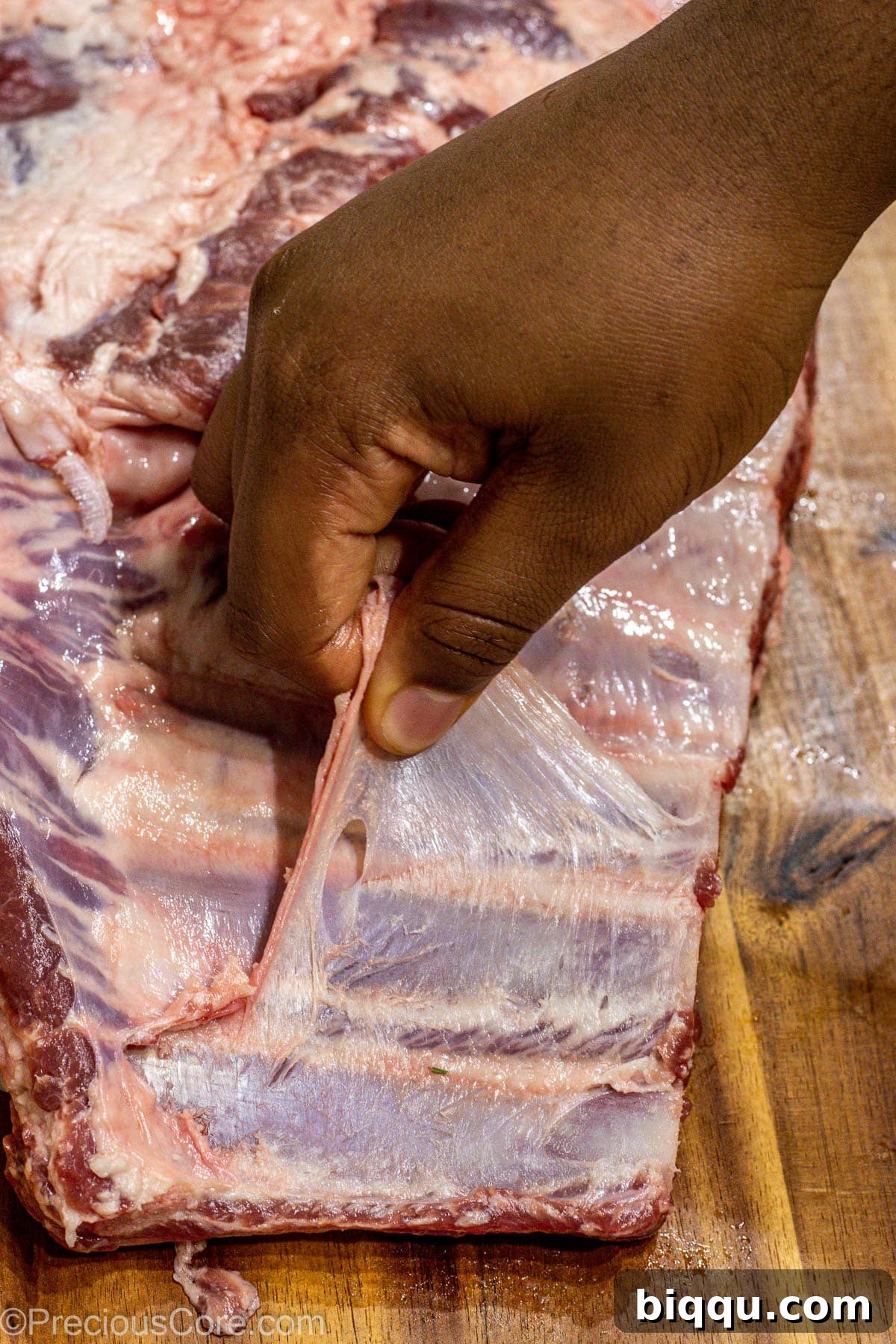 A hand expertly pulling the silver skin membrane from the back of a rack of spare ribs on a cutting board, revealing the meaty underside.