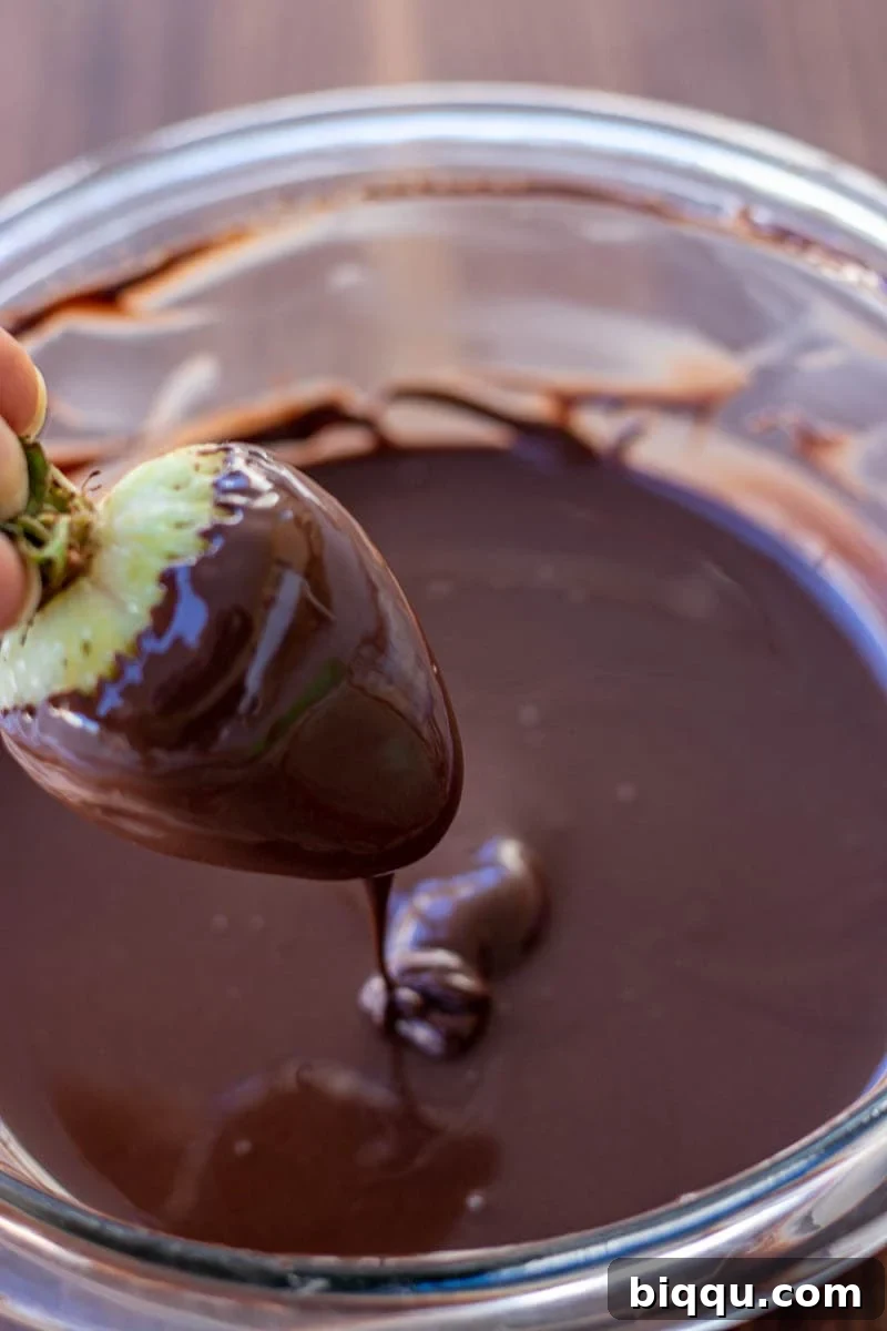 A hand gently holds a perfectly ripe strawberry, half-dipped in a pool of smooth, melted dark chocolate, capturing the moment of transformation.