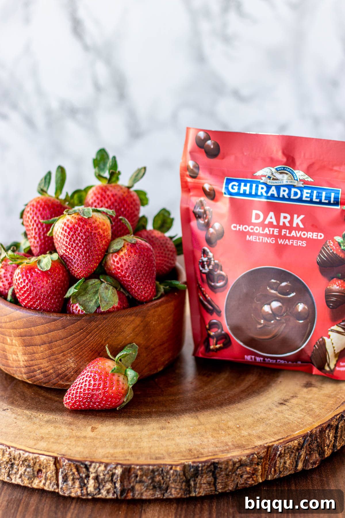 A bowl brimming with fresh, ripe strawberries next to a bag of melting chocolate wafers, ready for the dipping process to begin.
