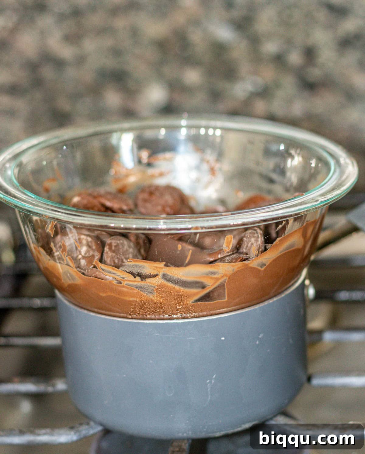 A glass mixing bowl filled with chocolate wafers sits atop a saucepan of simmering water, illustrating the double boiler method for melting chocolate.