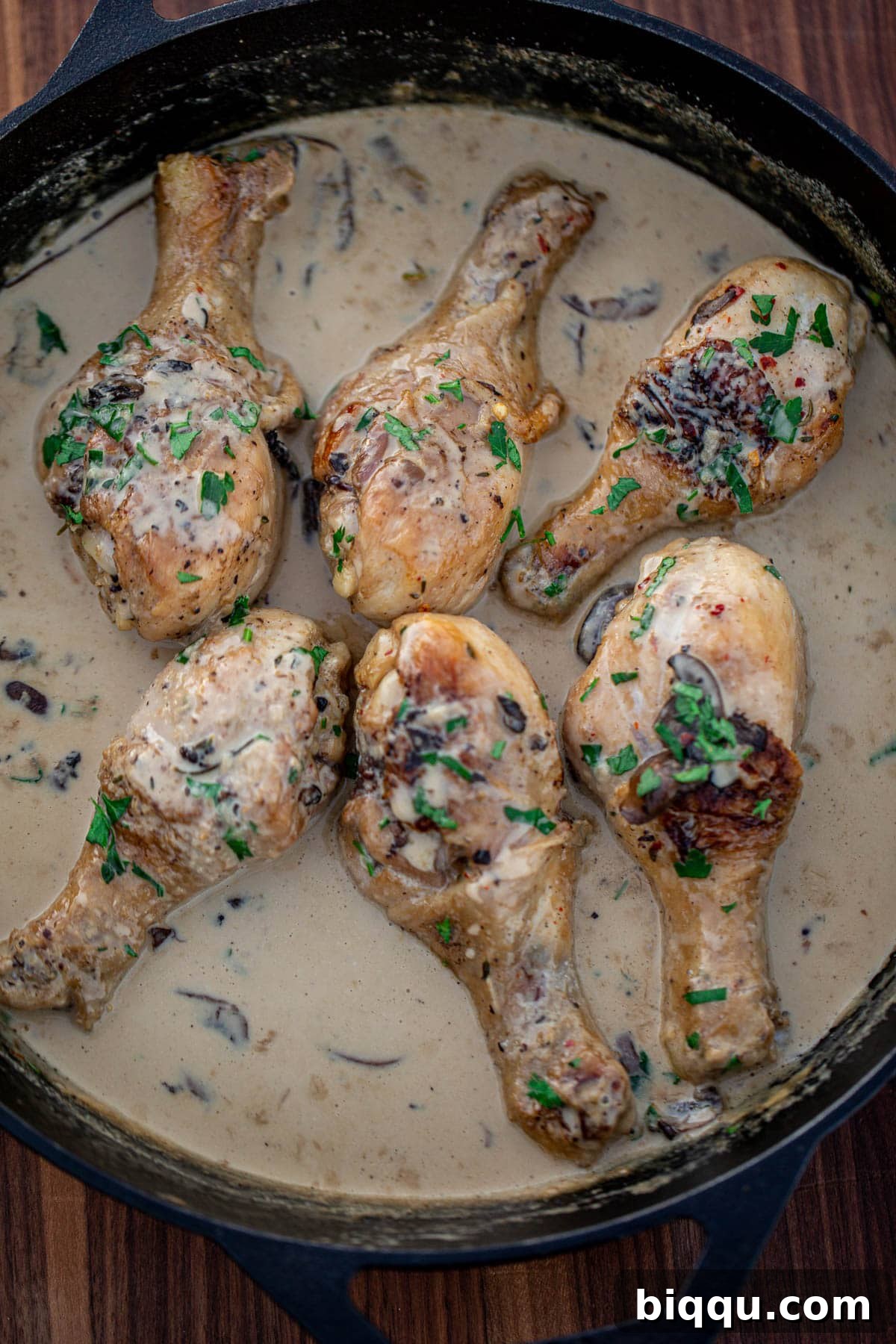 Golden-brown chicken drumsticks generously coated in a luscious creamy mushroom sauce, served directly from a cast iron skillet, garnished with fresh herbs.