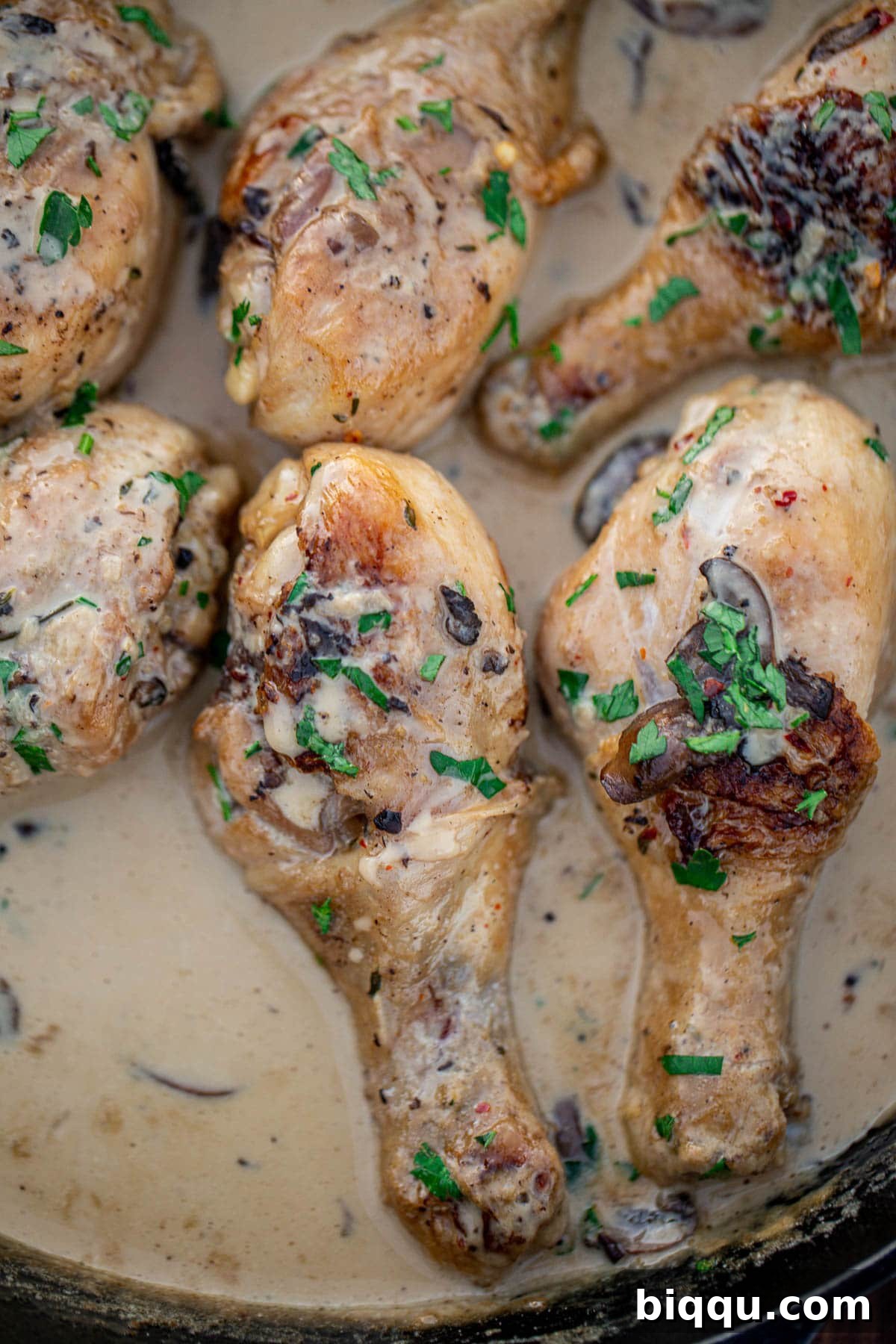 Close-up shot of succulent chicken drumsticks in creamy sauce, highlighting the texture of the sauce and mushrooms, garnished with fresh herbs.