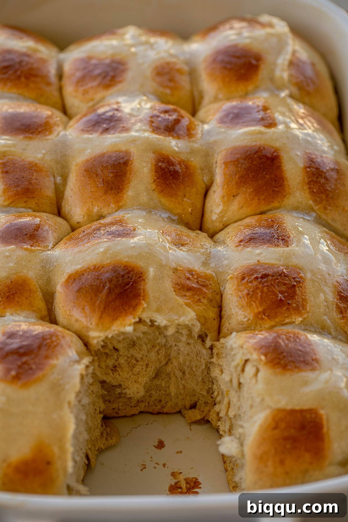 A baking dish filled with golden-brown Hot Cross Buns, with one bun removed, showing its soft interior and perfect cross.