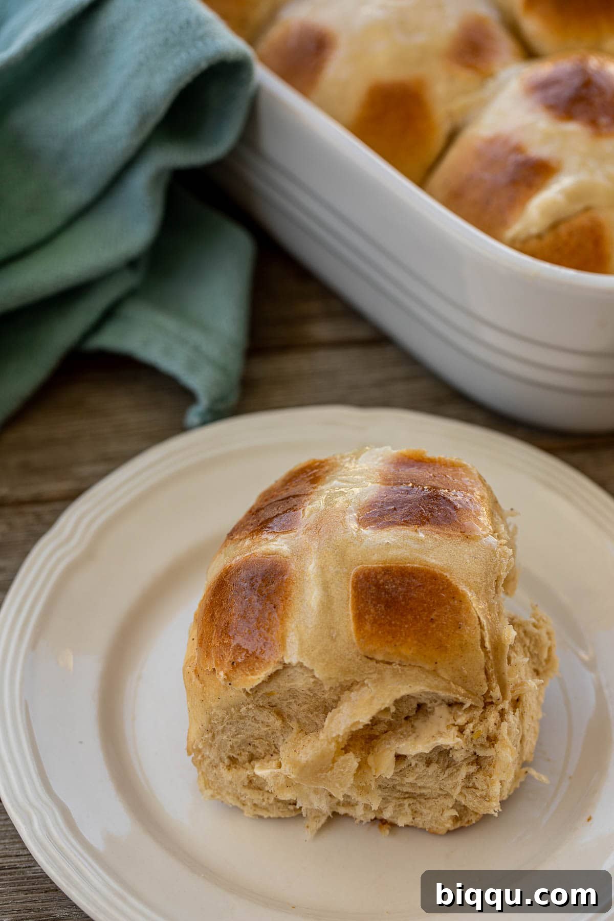 A single sweet spiced Hot Cross Bun resting on a white plate, showcasing its fluffy texture and golden crust.