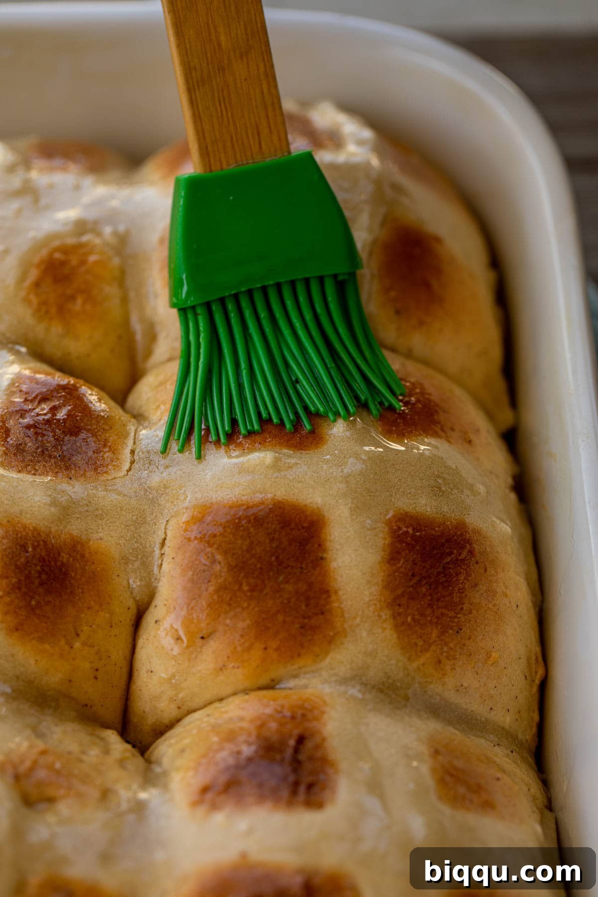 A close-up shot of a pastry brush applying a shiny lemon glaze onto warm Hot Cross Buns in a baking dish.