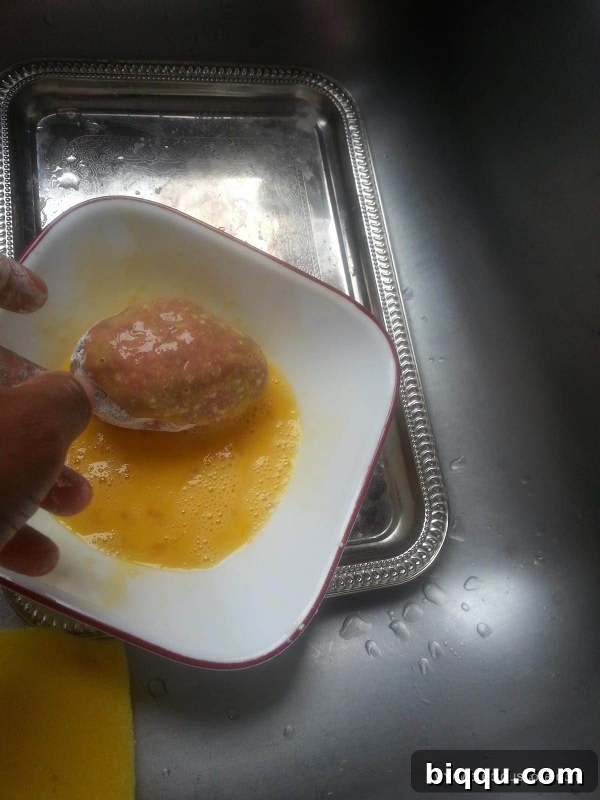Scotch Egg coated in flour, now being carefully dipped into a bowl of beaten egg wash.
