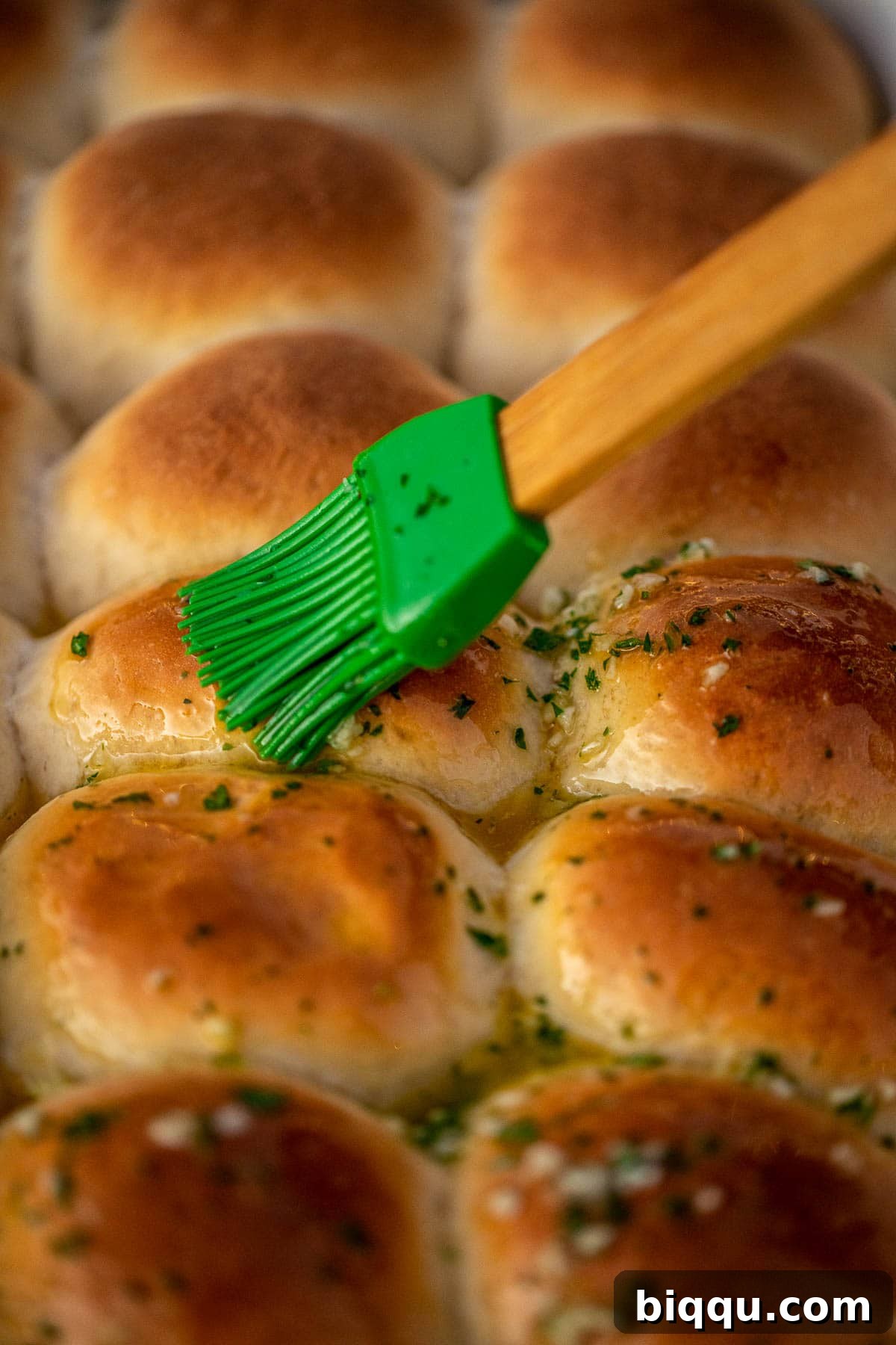 A hand brushing melted garlic butter over hot dinner rolls in a baking dish.