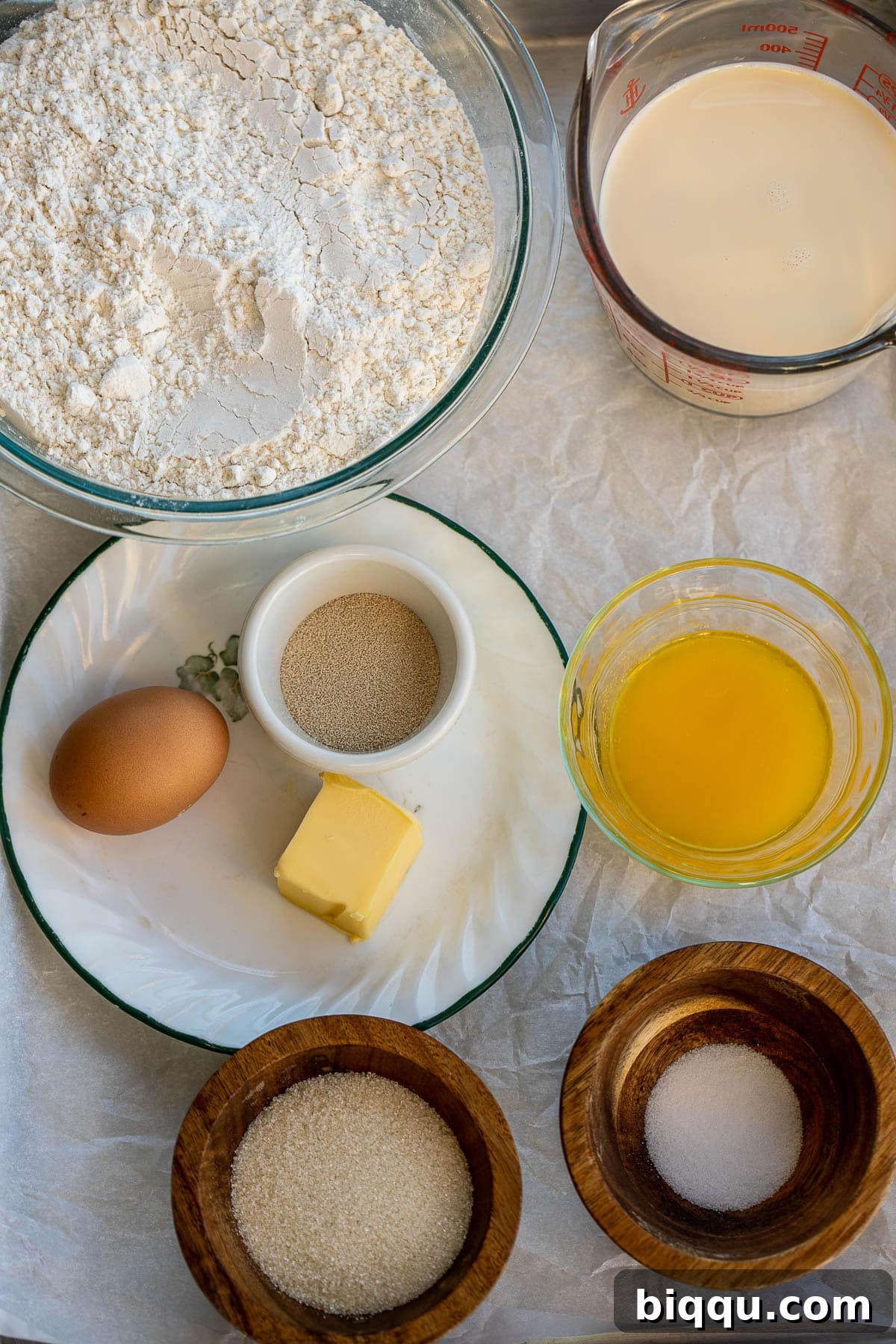 Various ingredients for making dinner rolls laid out on a kitchen surface, including flour, sugar, salt, butter, egg, and evaporated milk.