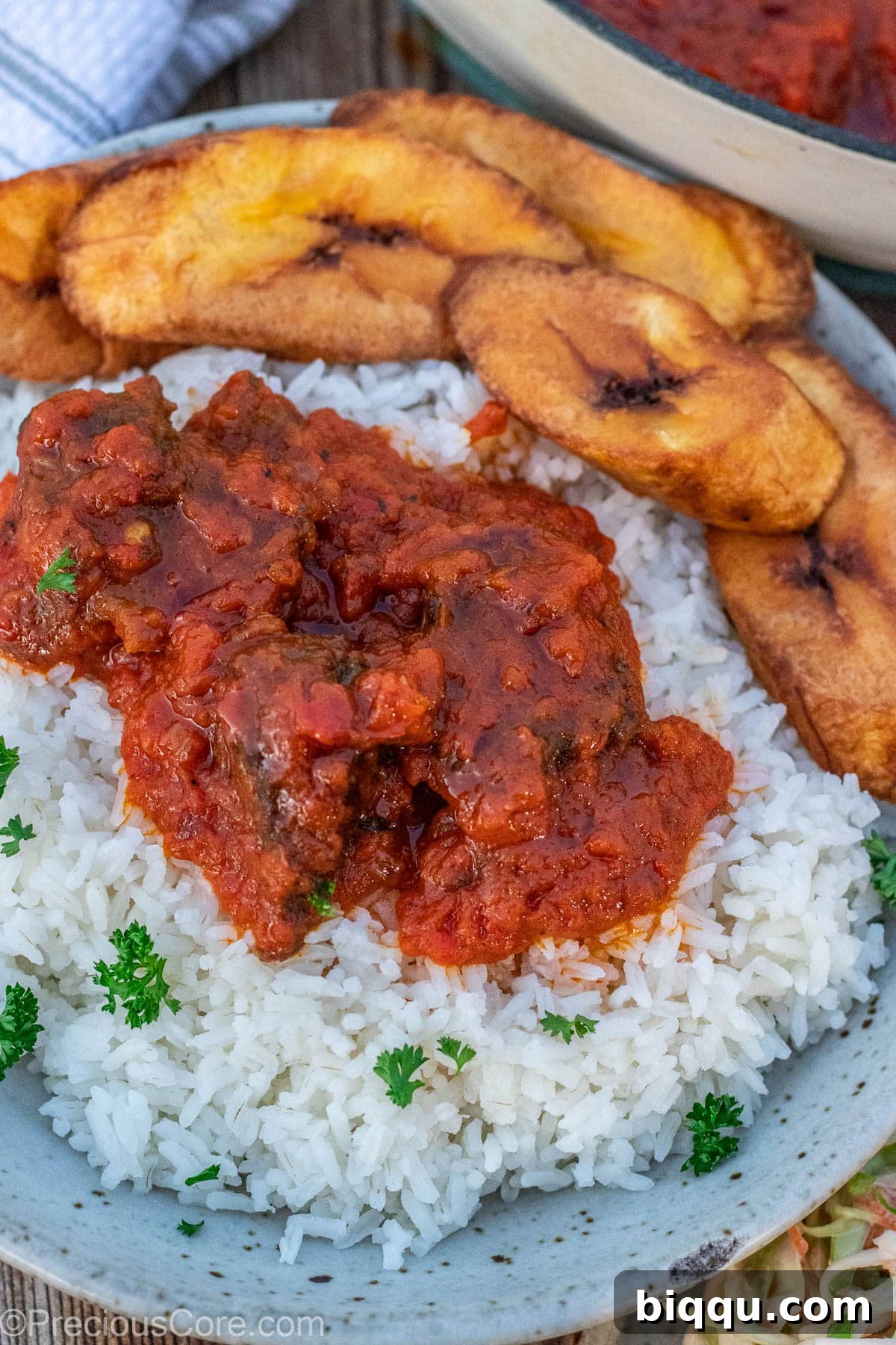 A bowl of rice, topped generously with tomato beef stew and accompanied by sweet fried plantains.