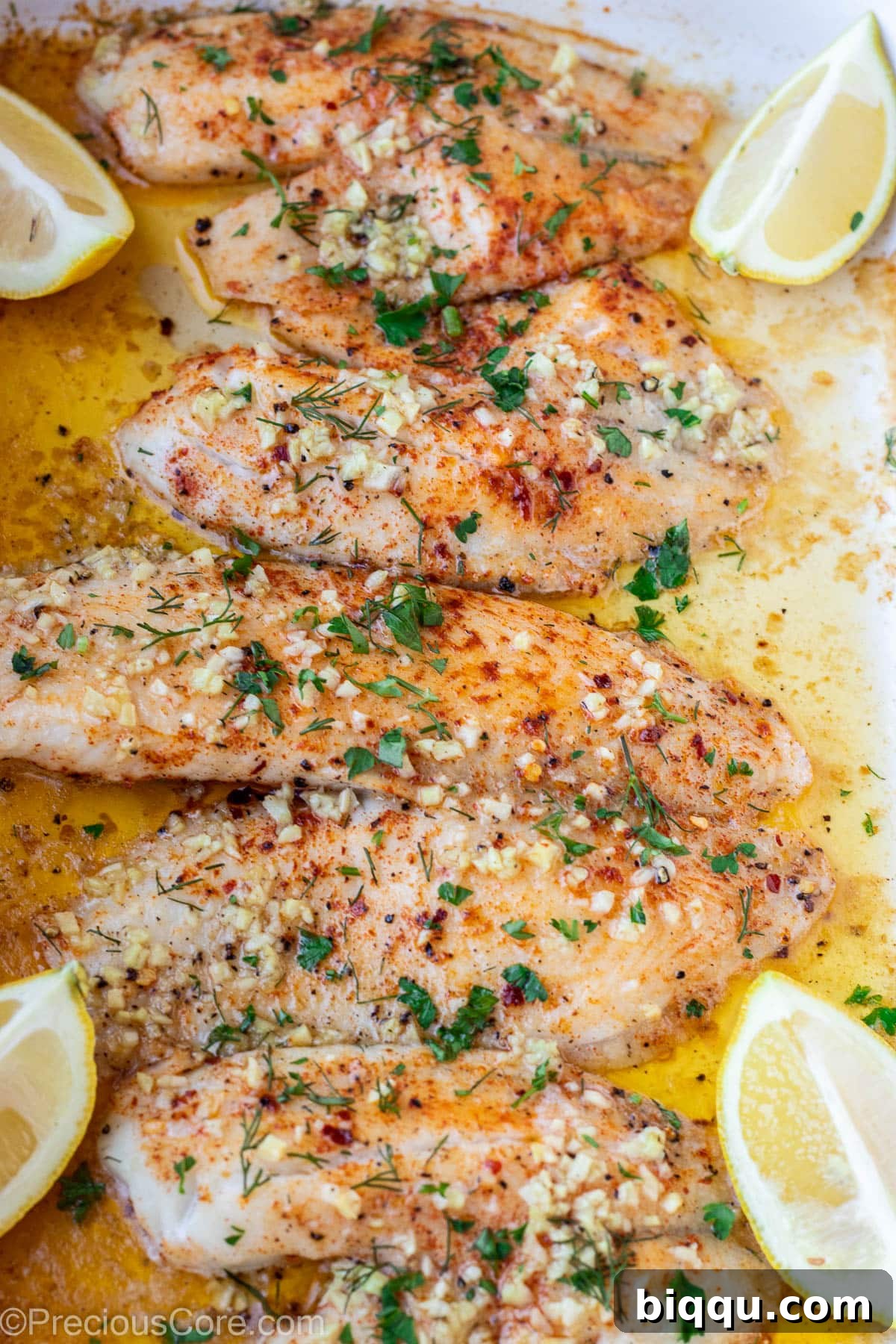 Four baked tilapia fillets glistening with garlic butter in a baking pan, garnished with fresh parsley.