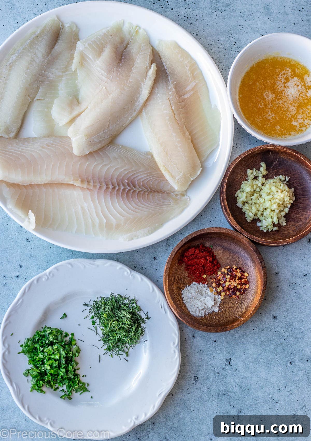 A collection of fresh ingredients for garlic butter tilapia including fillets, butter, garlic, and spices, laid out on a kitchen slab.