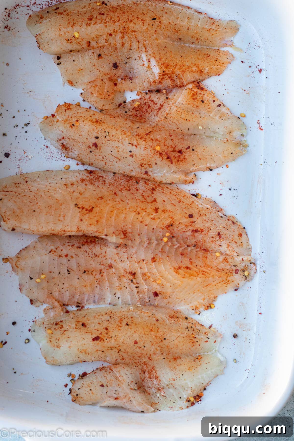 Tilapia fillets expertly seasoned with a vibrant blend of spices in a white mixing bowl, ready for the next step.