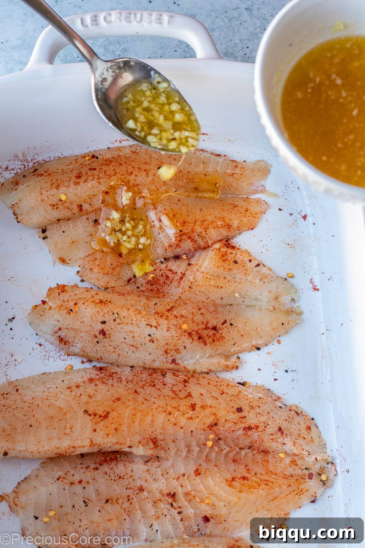 A spoon expertly drizzling rich, melted garlic butter sauce over seasoned raw tilapia fillets arranged in a baking dish.