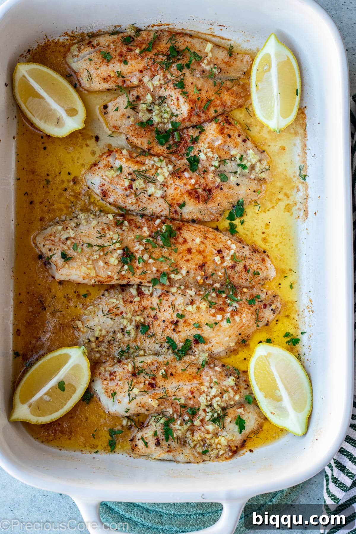 Four perfectly baked tilapia fillets in a glass baking dish, golden and flaky, fresh from the oven and garnished with herbs.