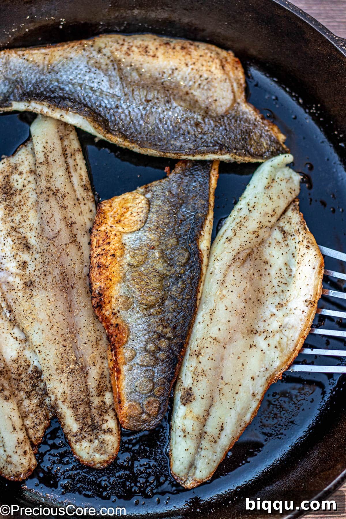A fish spatula flipping a perfectly seared sea bass fillet in a cast iron pan.