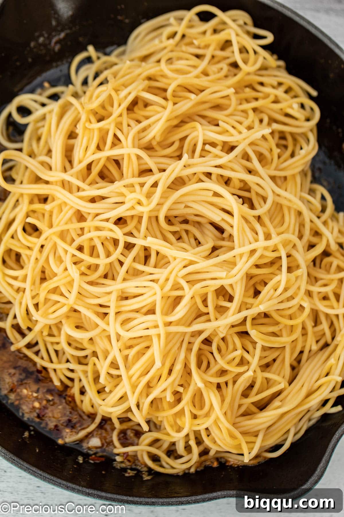 Cooked spaghetti being added to a cast iron pan containing the garlic butter sauce, ready to be tossed.