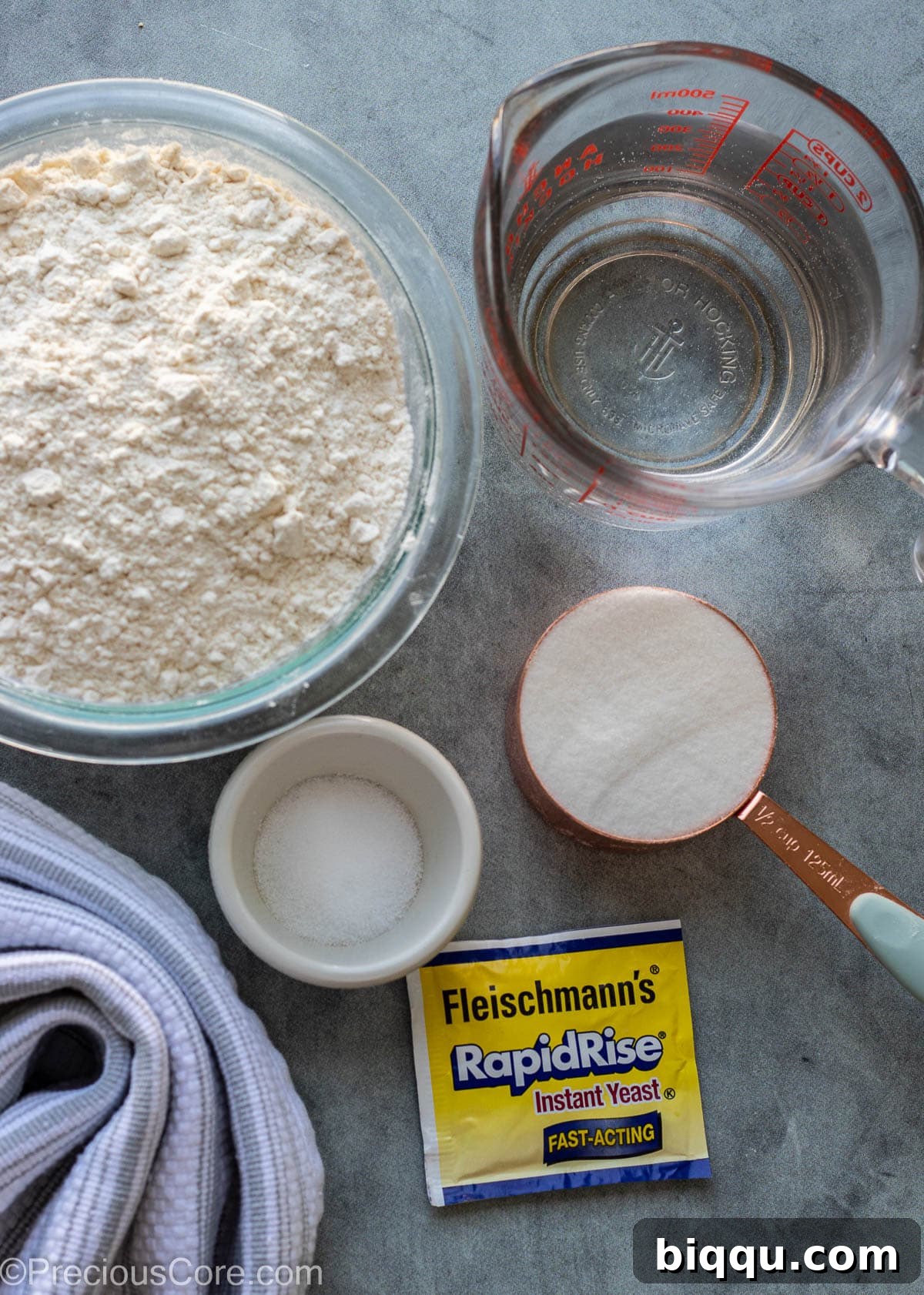 A clear glass bowl holding all-purpose flour, a measuring cup with sugar, active dry yeast, and salt, representing the core ingredients for Puff Puff.