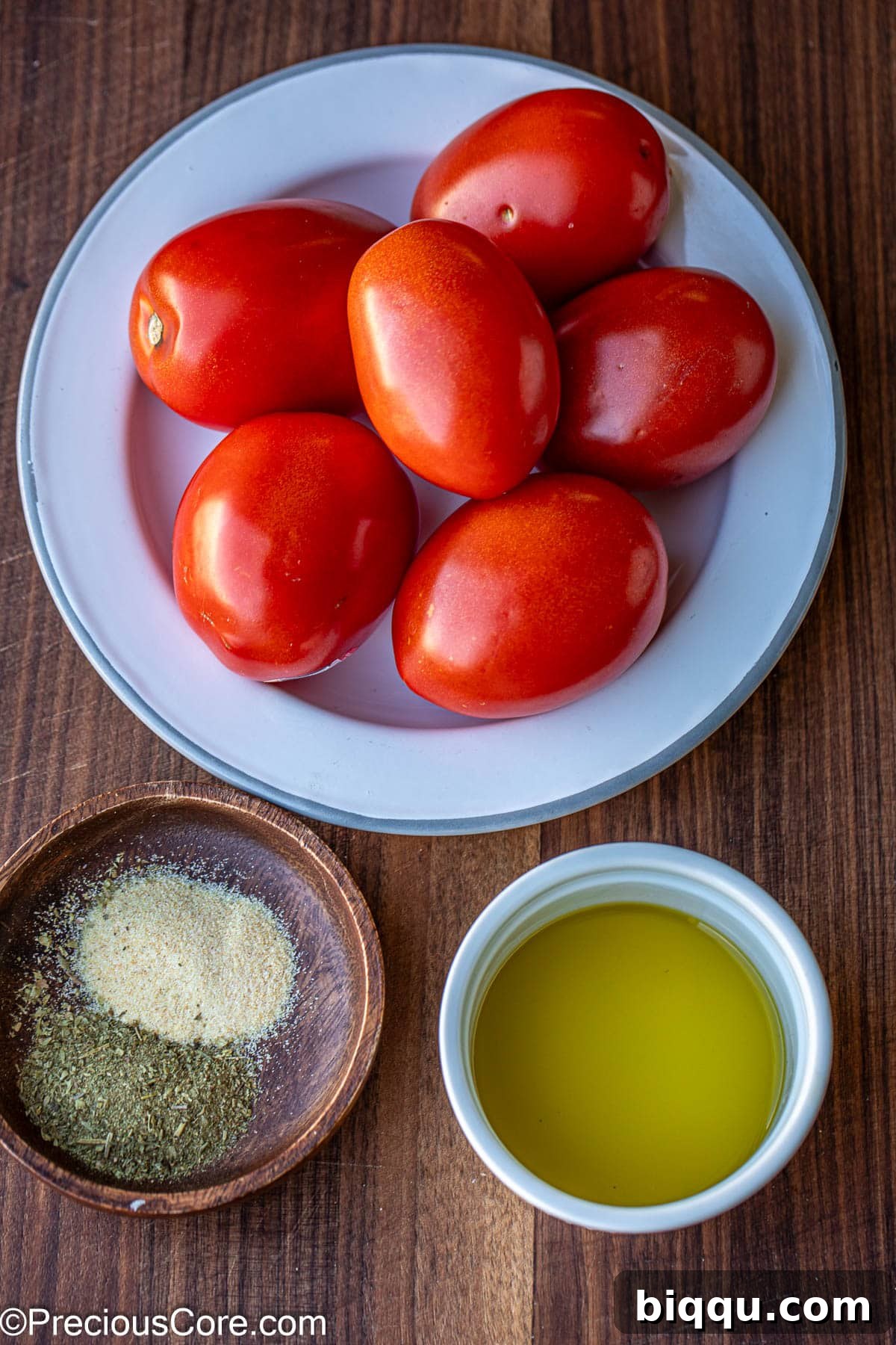 Fresh Roma tomatoes on a white plate, a wooden bowl with Italian spices, and a small bowl of olive oil.