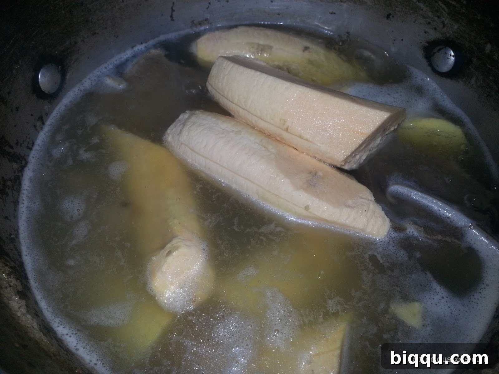 Unripe plantain chunks being added to a bubbling pot containing smoked meat and its rich broth.
