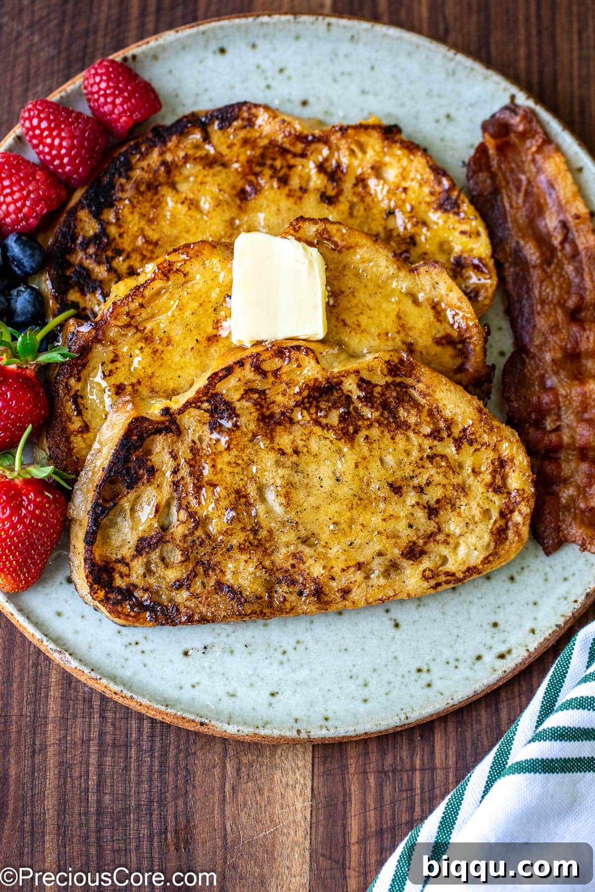 Three slices of perfectly cooked sourdough French toast on a plate, accompanied by fresh fruit and crispy bacon.