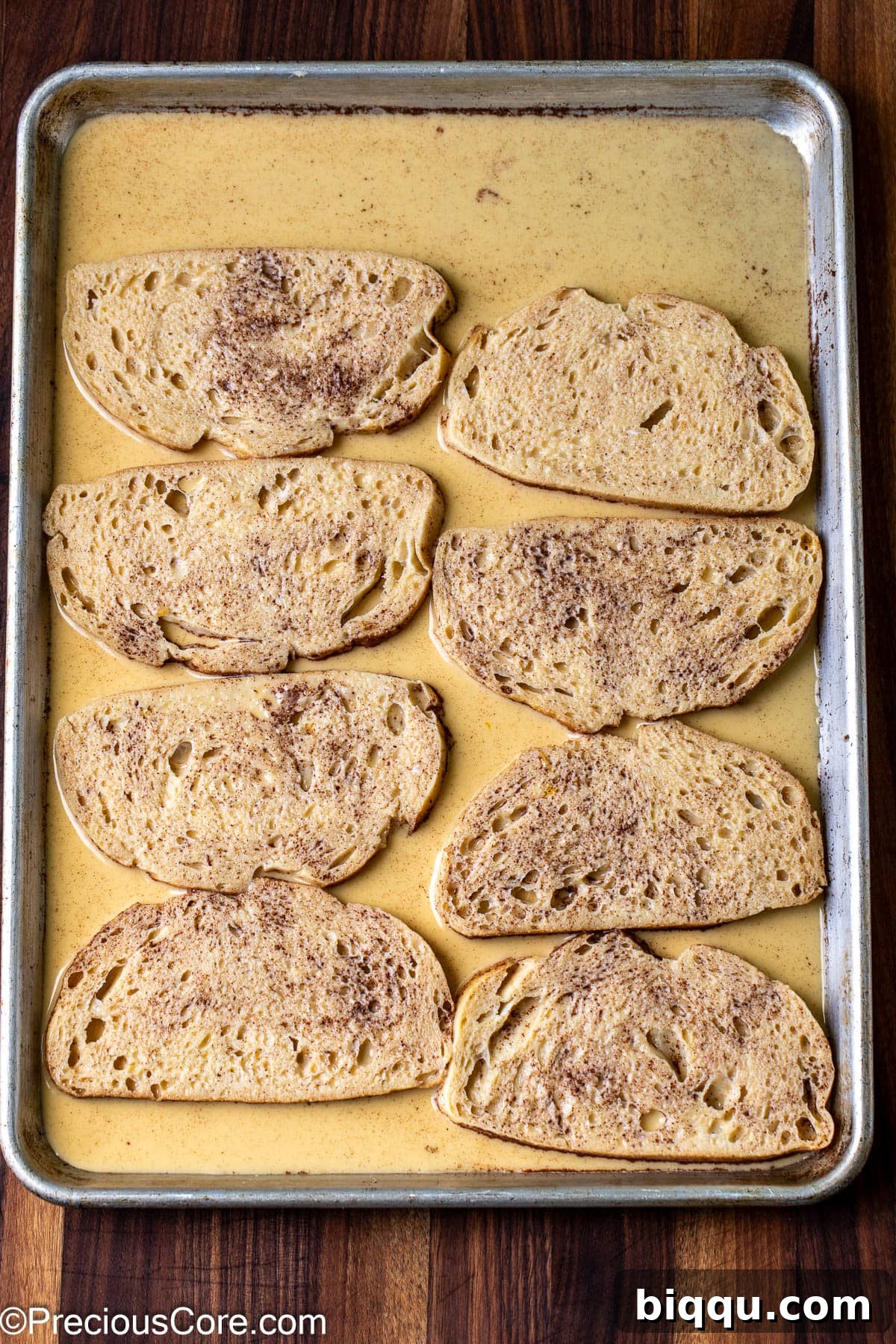 Slices of sourdough bread soaking in the rich custard mixture on a flat baking sheet.