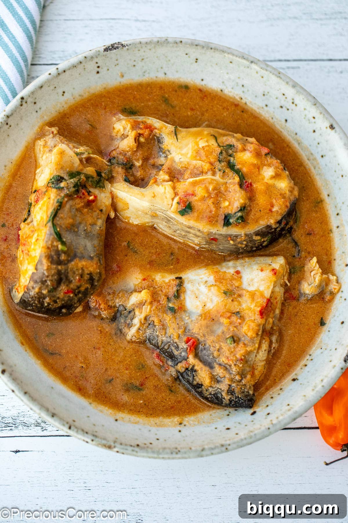 A vibrant bowl of Catfish Pepper Soup, featuring three succulent steaks of catfish in a rich, amber broth, garnished with fresh herbs.