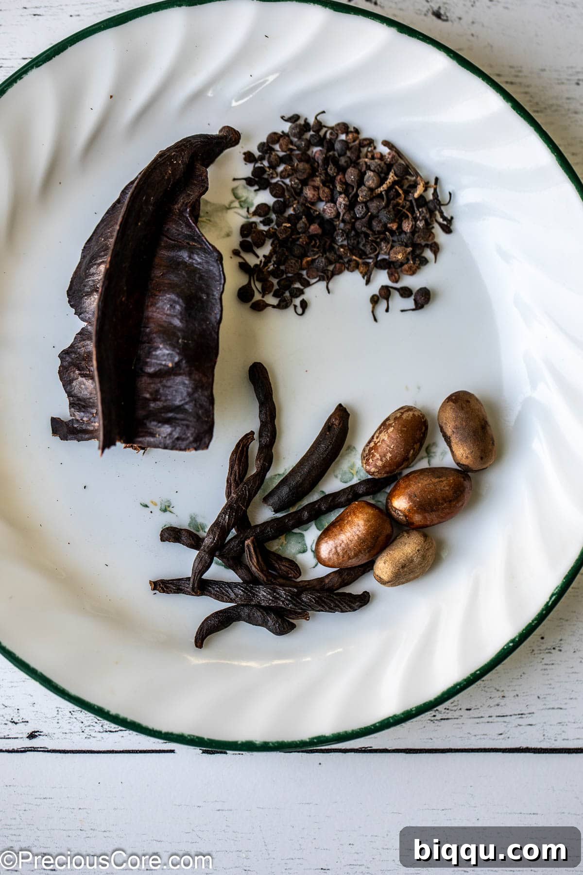 A collection of traditional West African pepper soup spices including Aidan fruit, African black pepper, negro pepper, and African nutmeg, neatly arranged on a pristine white plate.