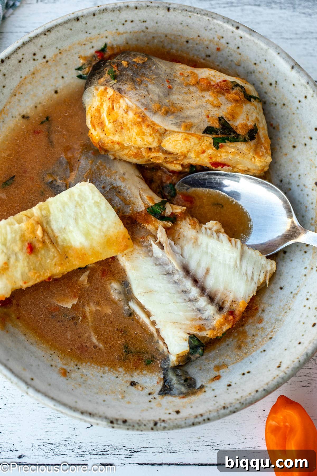 A close-up shot of a fish tail submerged in the spicy broth of pepper soup, served alongside boiled African white yam chunks, ready to be enjoyed.