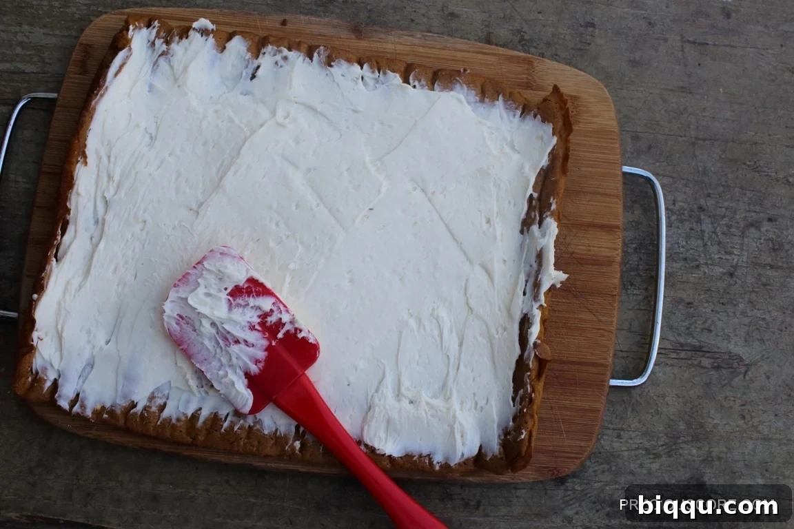 A red spatula spreading creamy cream cheese filling onto a flattened pumpkin cake.