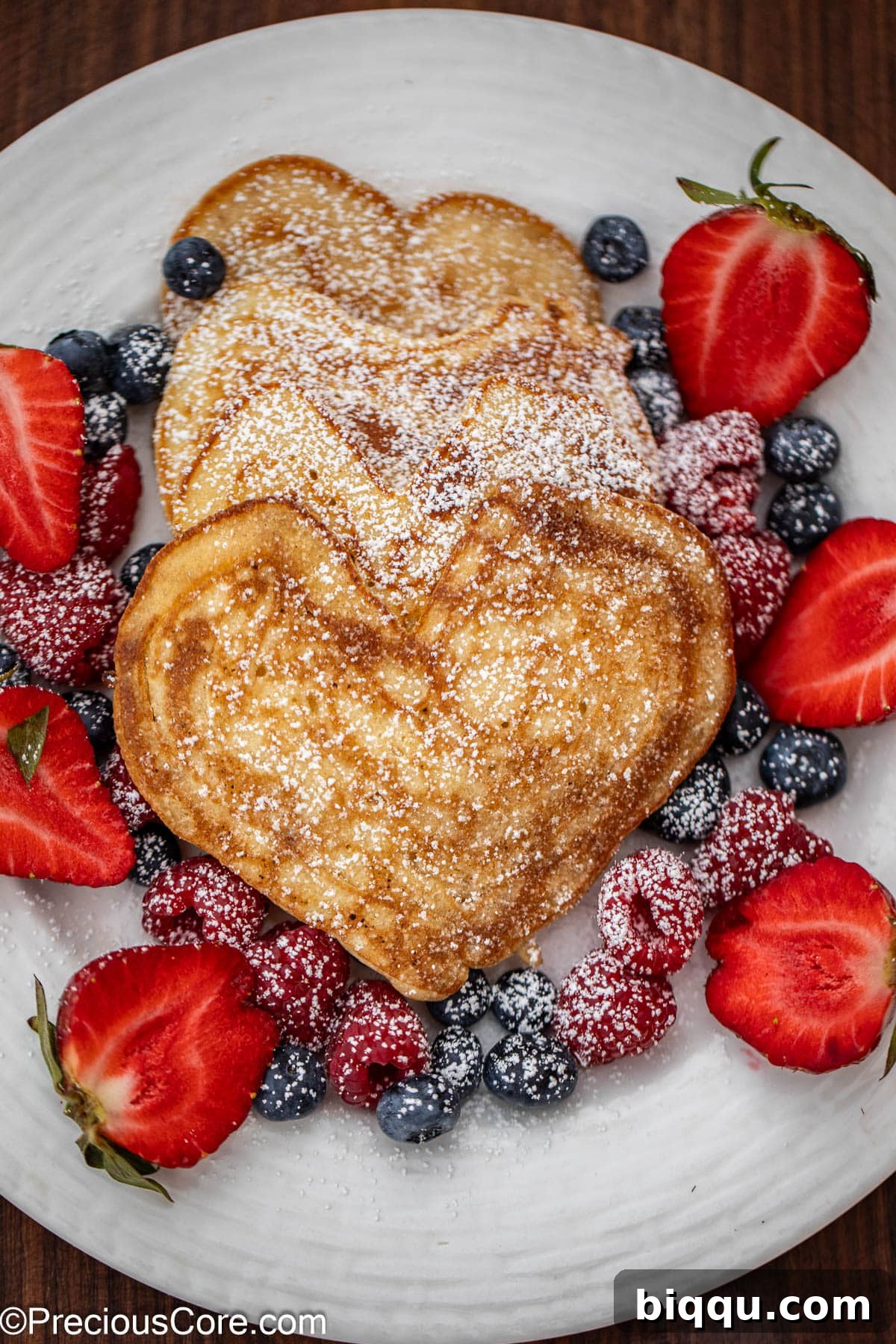 Heart shaped pancakes on a plate with fresh berries and powdered sugar, ready to be served.