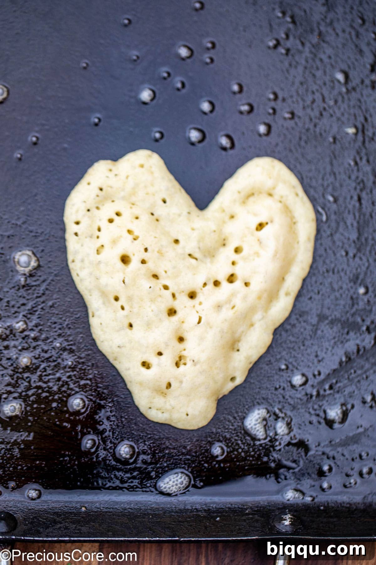 A heart-shaped pancake cooking on a griddle, showing the outline and filled center.