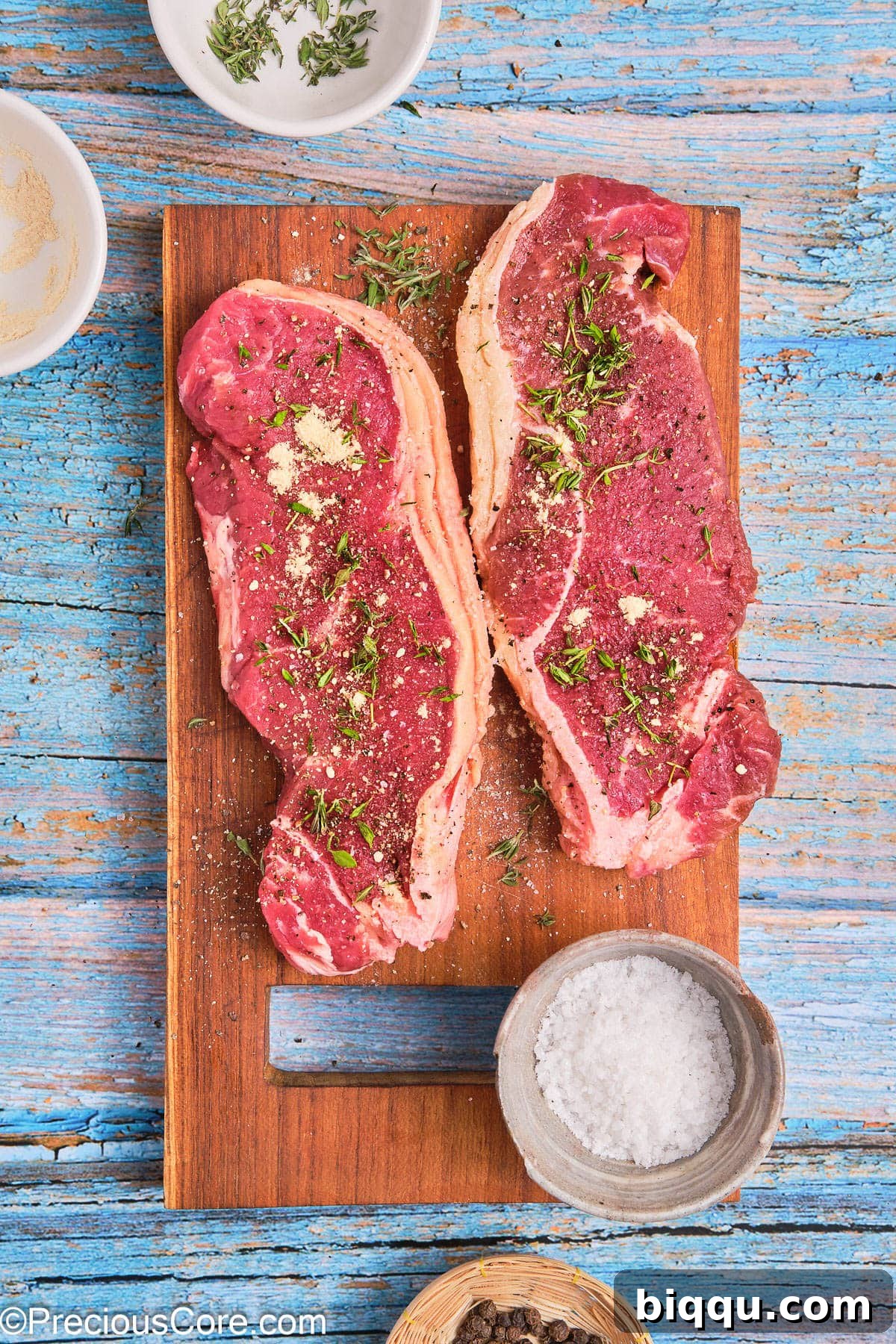 Two ribeye steaks, beautifully seasoned with a mix of herbs and spices, on a cutting board.