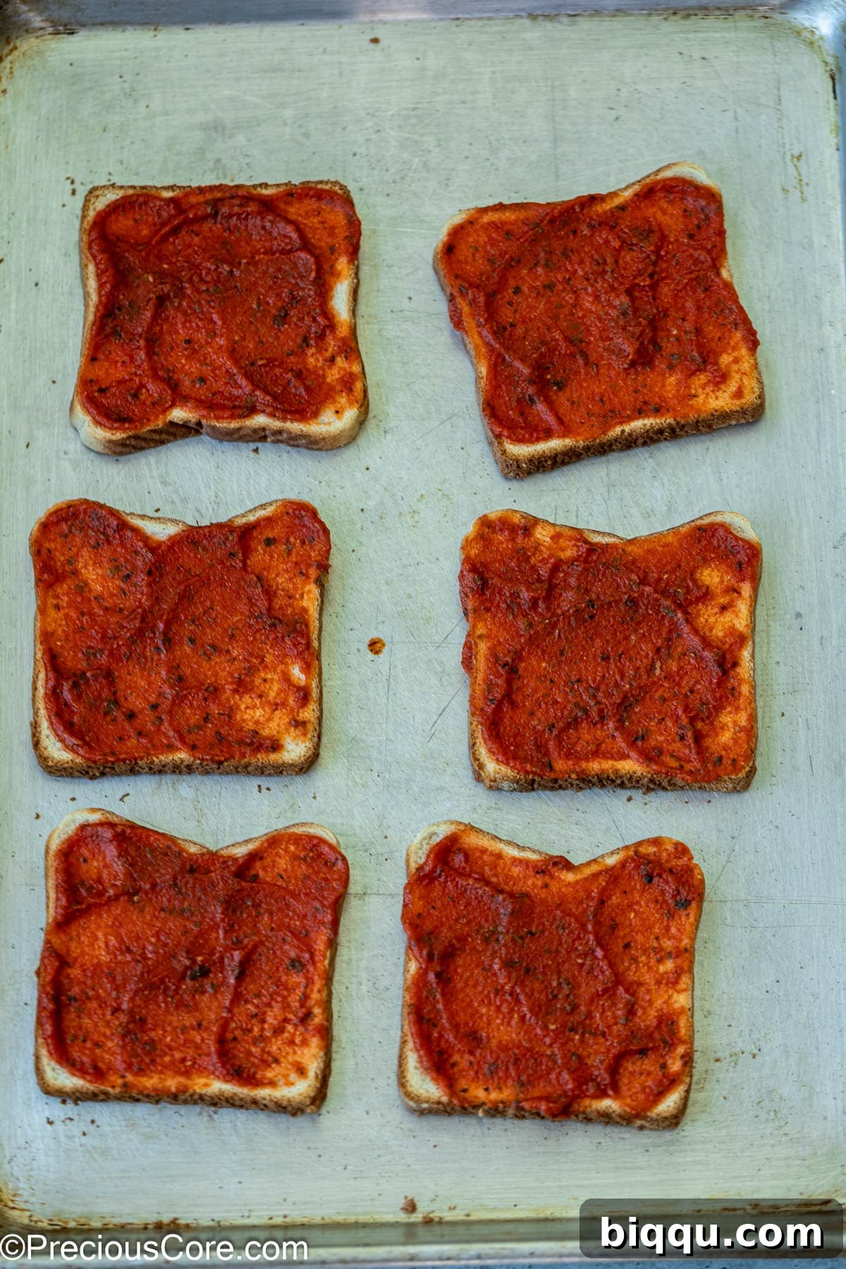 Pizza sauce being spread generously on lightly toasted bread slices.