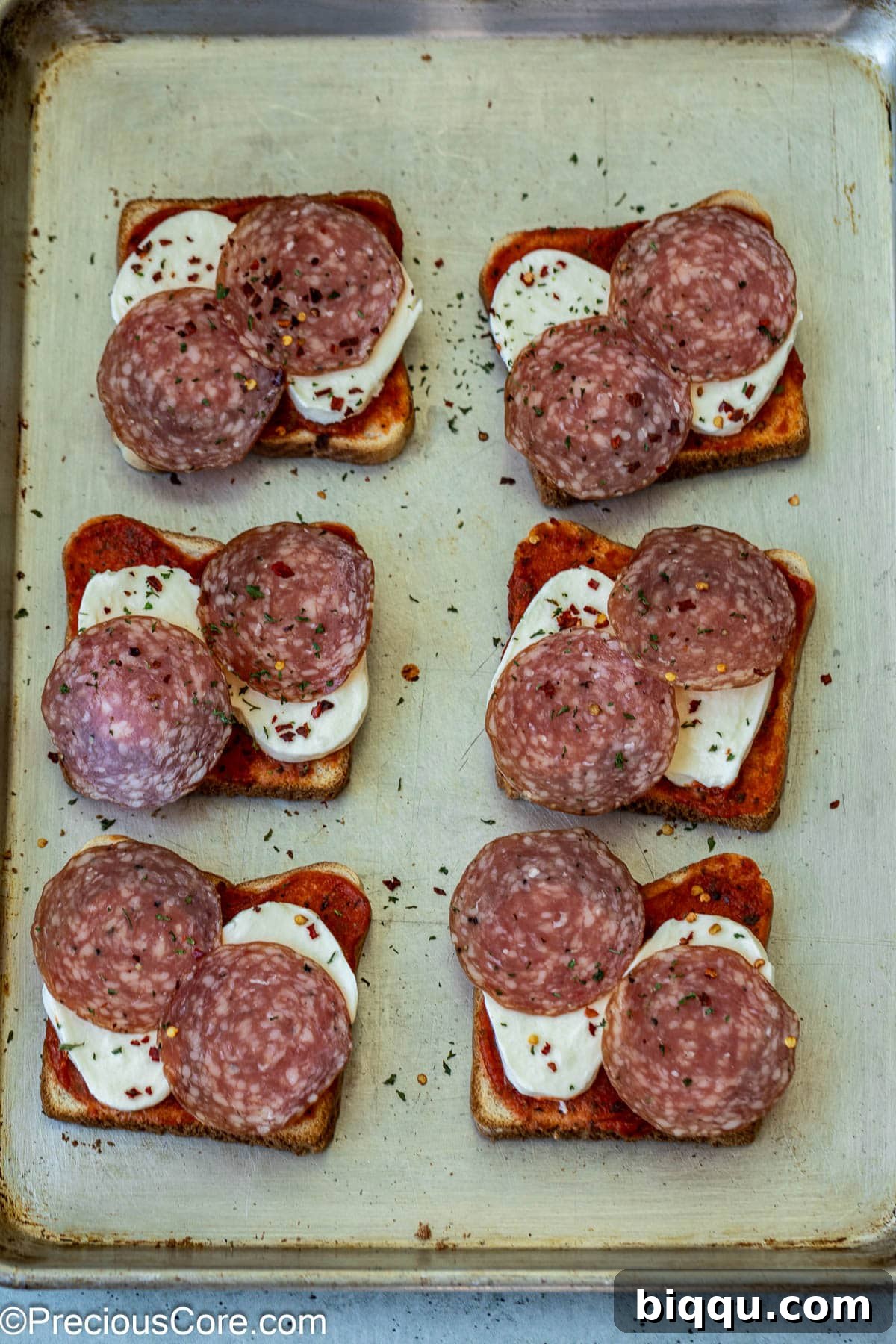 Unbaked pizza toasts topped with salami, parsley flakes, and red pepper flakes, ready for the oven.