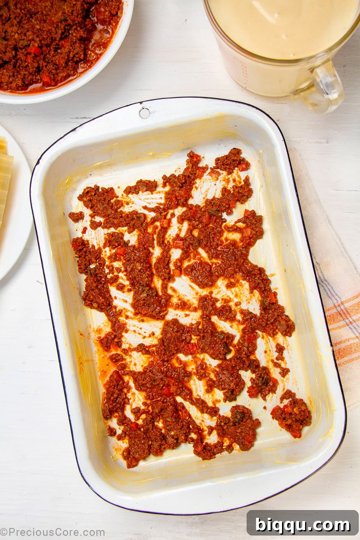A thin, even layer of rich meat sauce spread across the bottom of a baking dish, preparing for the first layer of pasta.