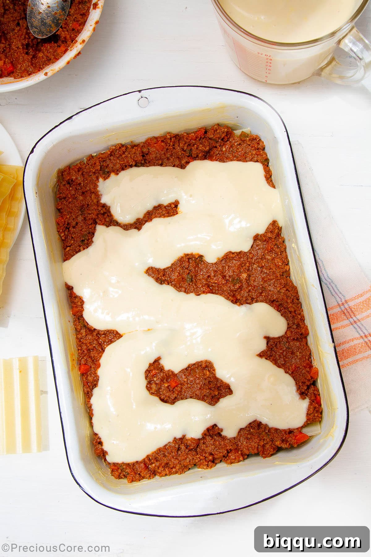 A smooth, white layer of béchamel sauce spread over the meat sauce and pasta in a baking dish, ready for the next layer.