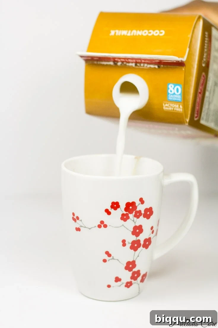 A carton pouring creamy coconut milk into a floral mug for a dairy-free hot chocolate.