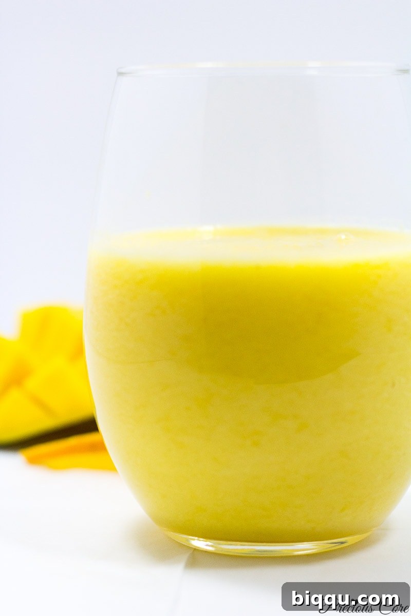 A vibrant yellow pineapple mango smoothie, garnished with a slice of fresh pineapple, served in a small clear glass on a light background.