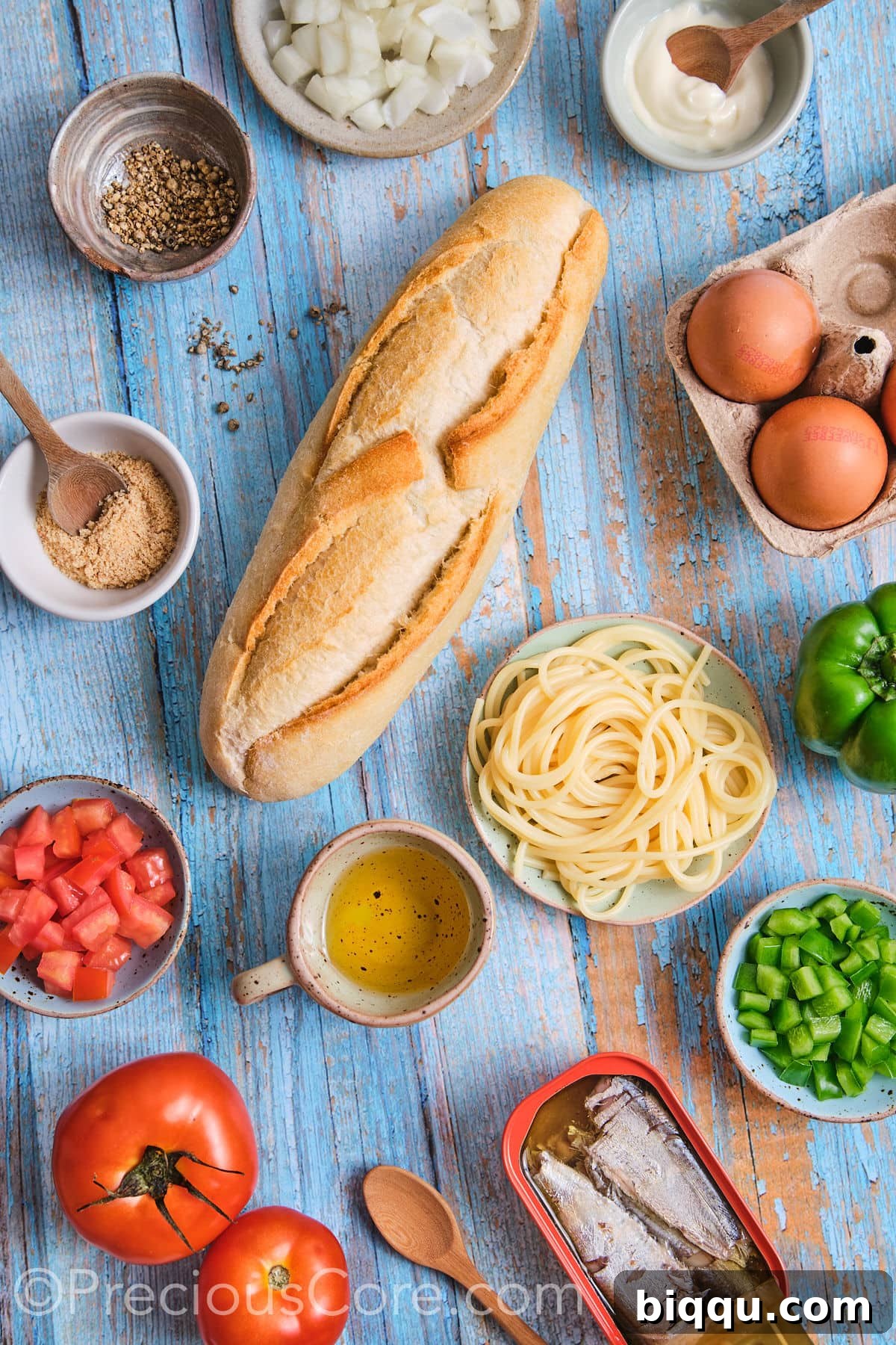 A flat lay photograph showcasing all the fresh and simple ingredients laid out for a homemade spaghetti omelette sandwich, including eggs, vegetables, spaghetti, and a baguette.