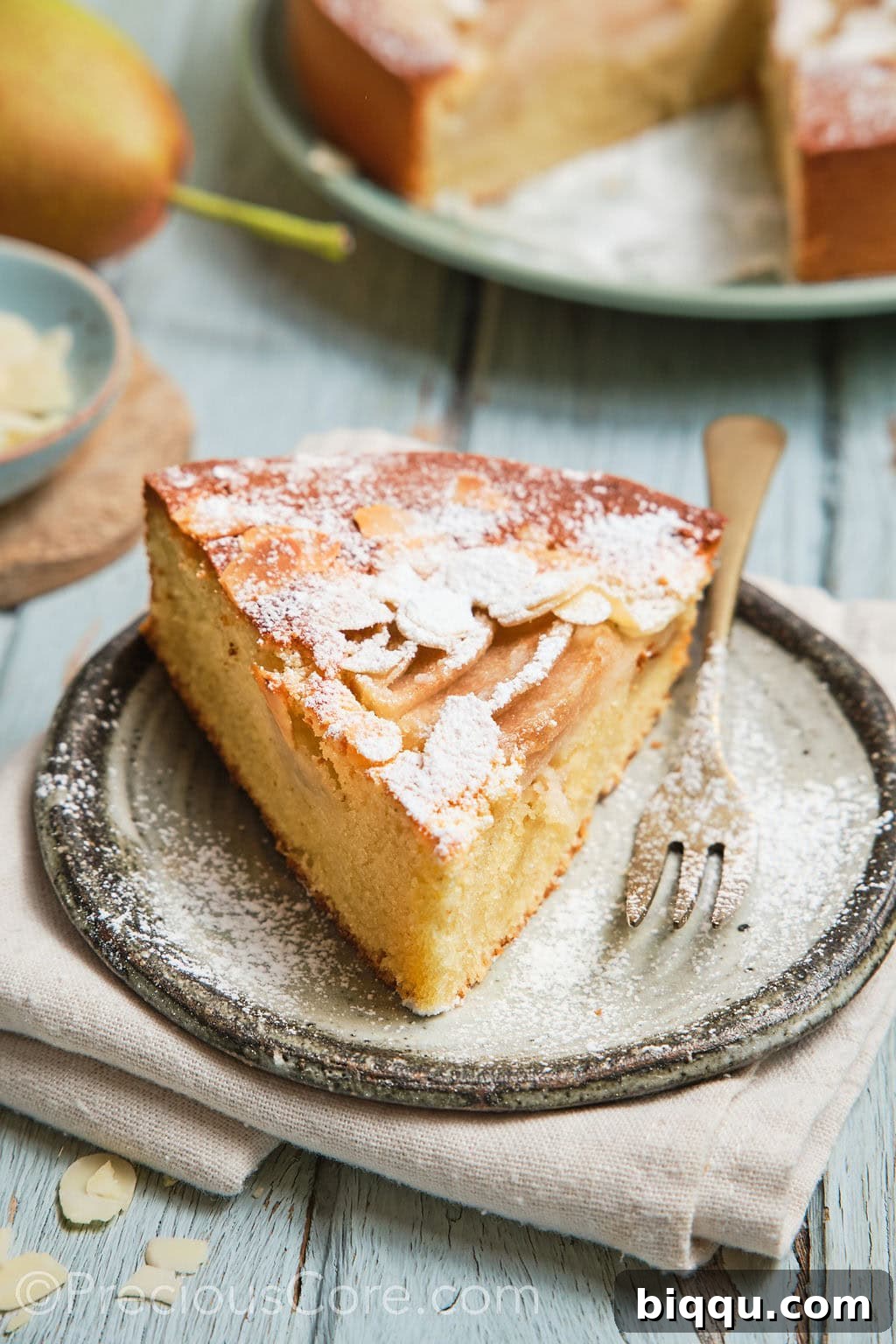 A slice of pear almond cake, dusted with icing sugar, served on a plate.