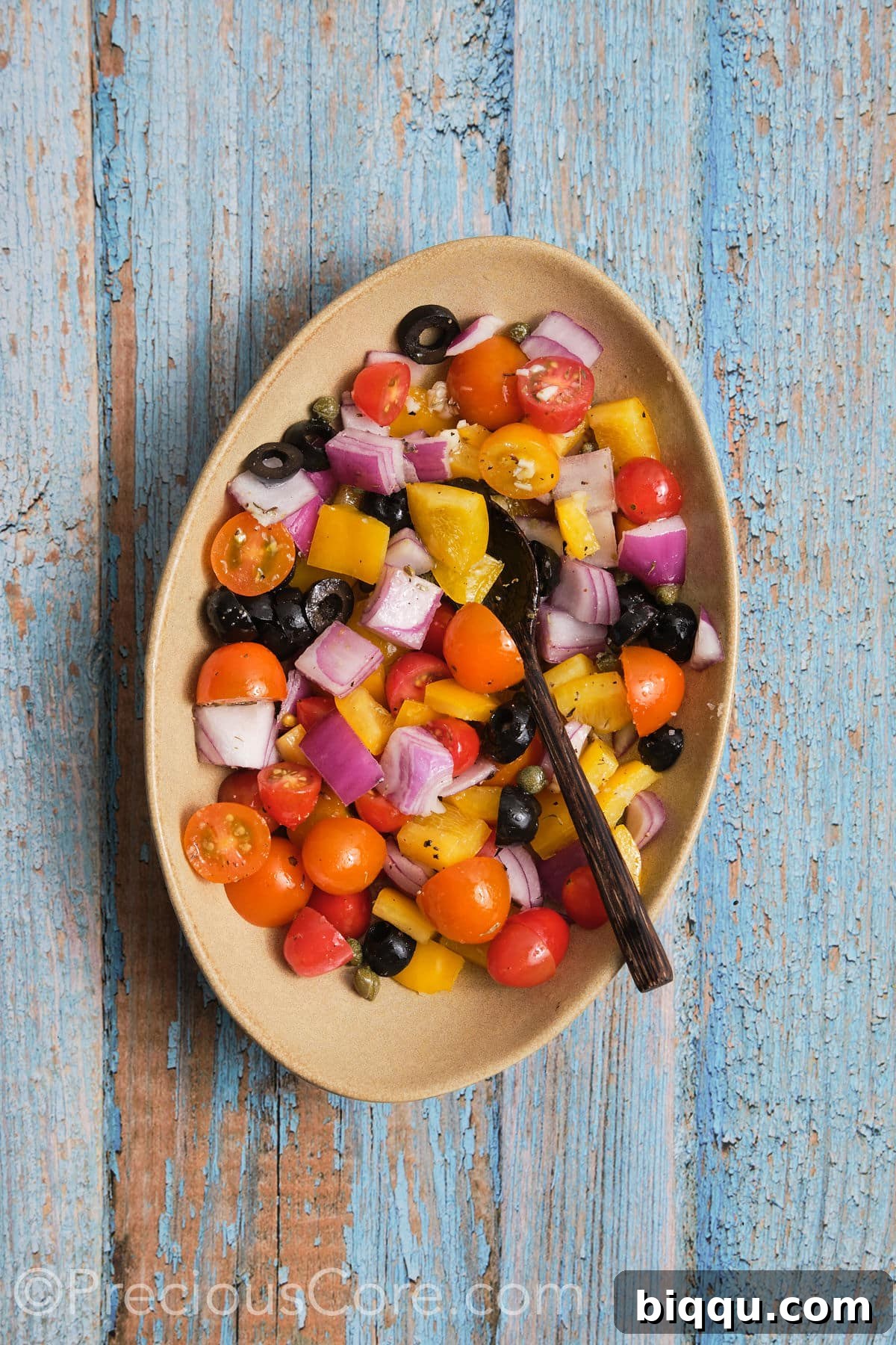 Diced Mediterranean vegetables, including bell peppers, red onions, cherry tomatoes, black olives, and capers, tossed in olive oil and seasonings in a bowl.