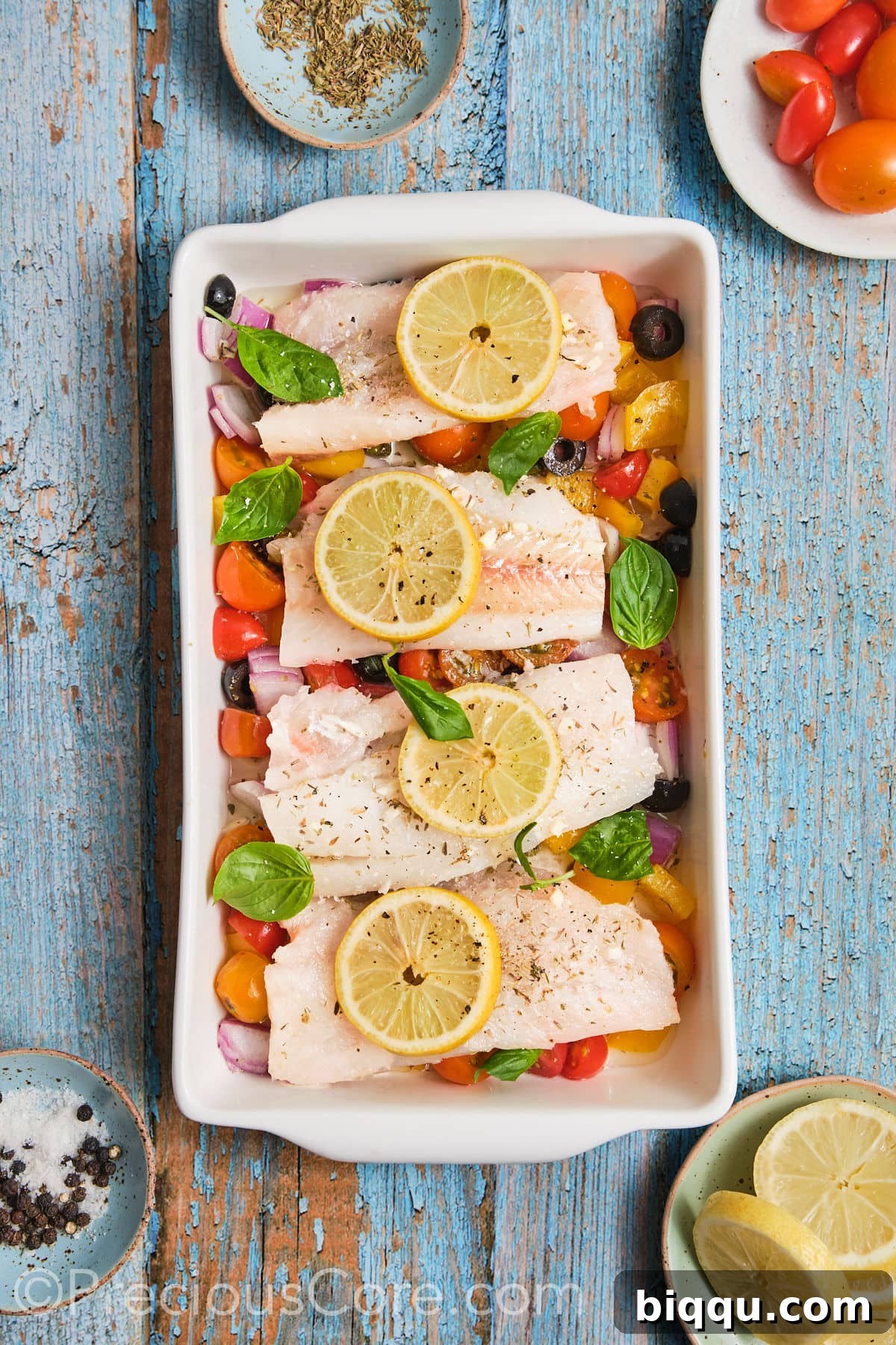 Baking dish filled with a bed of chopped vegetables, topped with marinated fish fillets and garnished with lemon slices, ready for the oven.