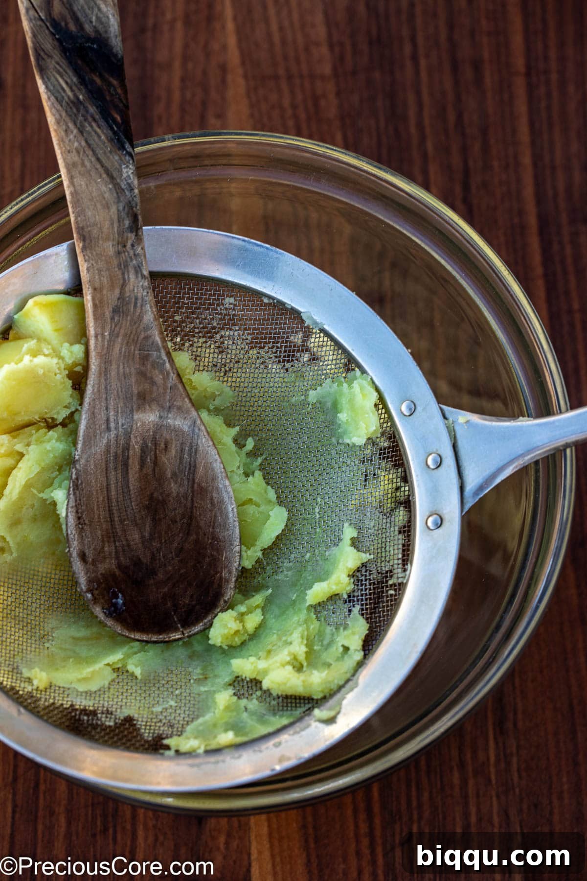 Hot, drained white sweet potatoes being pressed through a fine mesh sieve to achieve a smooth consistency.