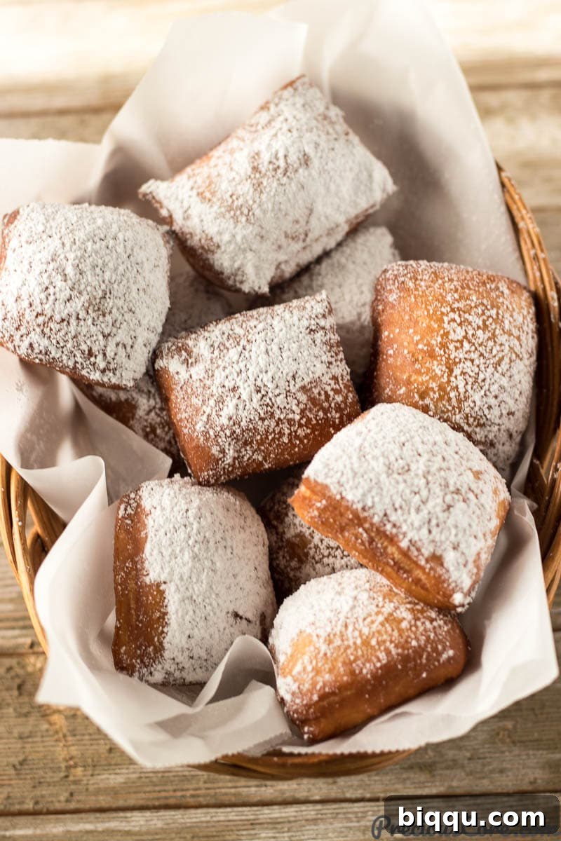 A close-up shot of several golden brown beignets piled in a rustic woven basket, covered in a snow-like layer of powdered sugar.