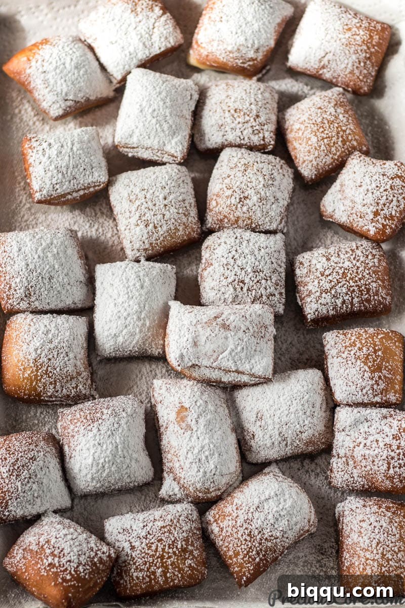 A delectable pile of freshly fried beignets generously dusted with powdered sugar, resting on a parchment-lined baking sheet, glistening under the light.