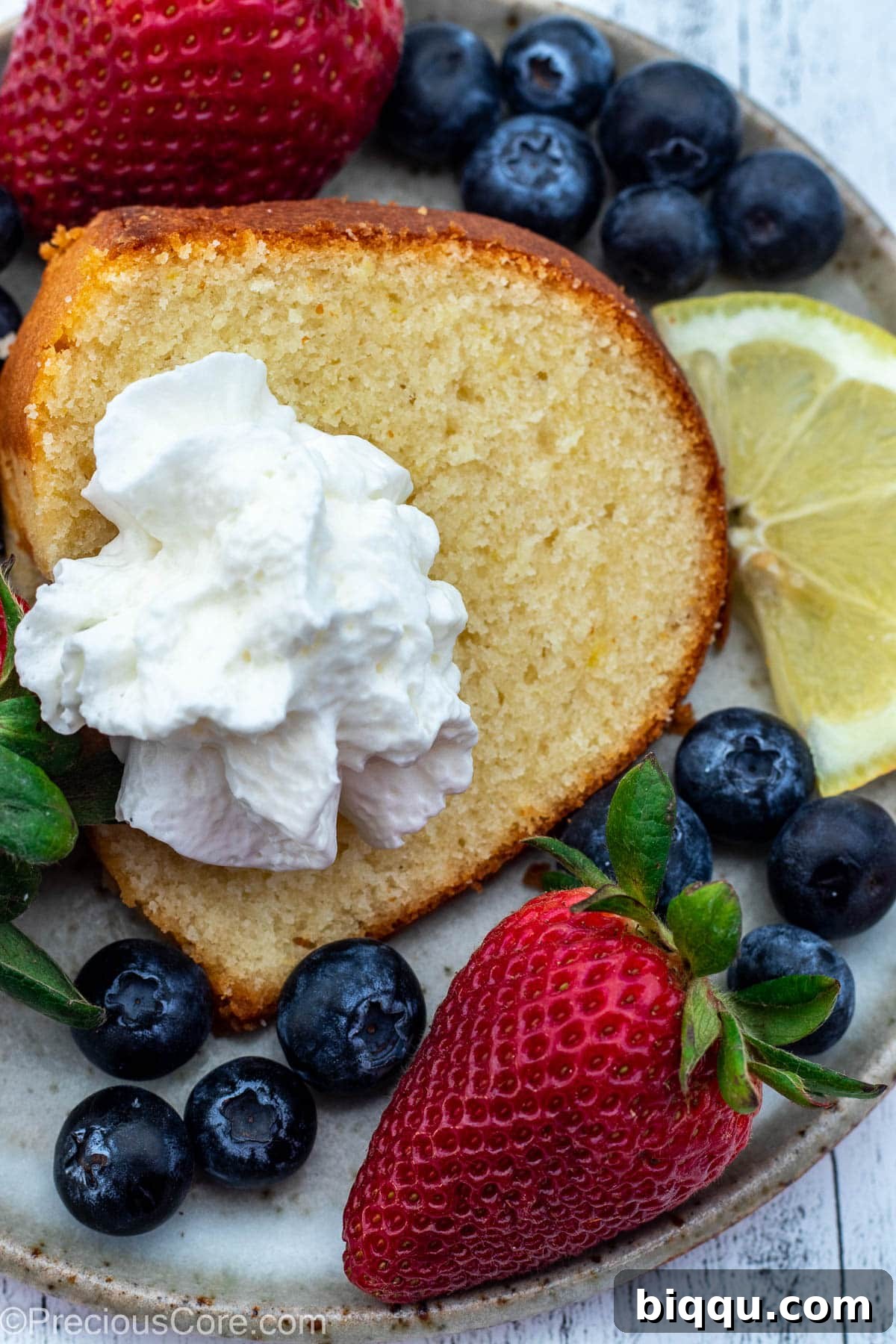 A slice of lemon pound cake on a plate, topped with whipped cream and surrounded by berries.